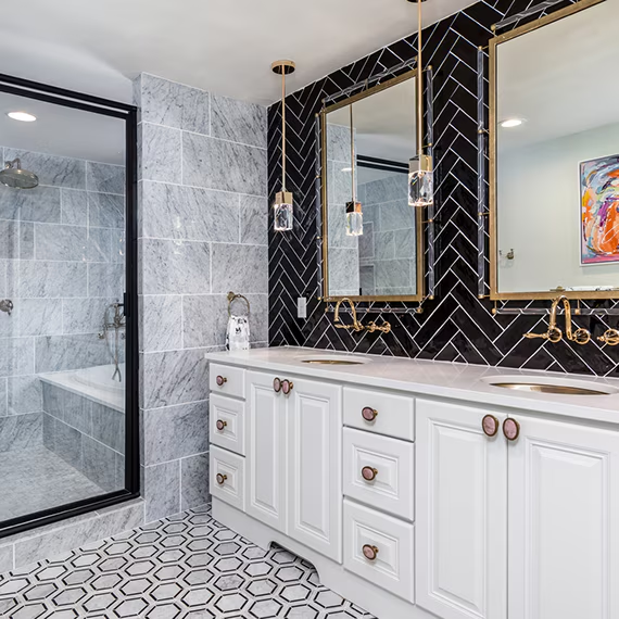 Modern bathroom with white vanity, gold hardware, black herringbone tile wall, marble-look shower tiles, and hexagonal patterned flooring